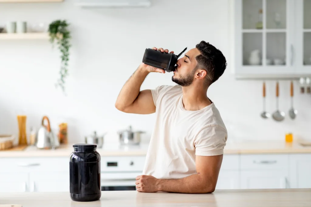 Hombre bebiendo un batido de un shaker en la cocina, junto a un bote de suplemento
