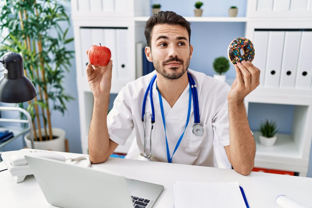 young hispanic dietitian man holding doughnut apple smiling looking side staring away thinking