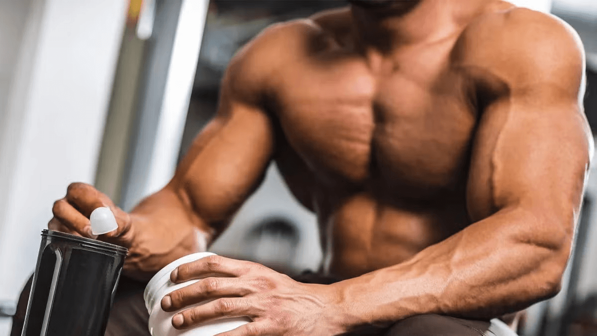 Hombre musculado sentado en un gimnasio abriendo un bote de suplemento junto a un vaso mezclador