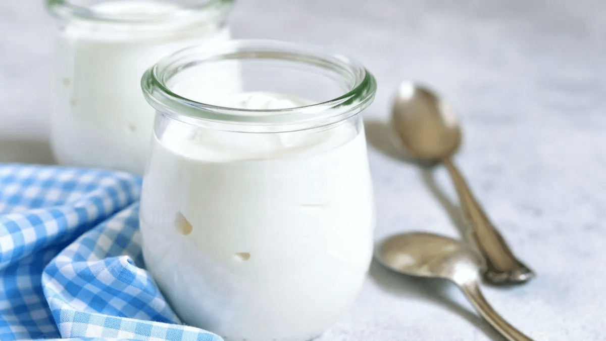 Dos tarrinas de cristal con yogur natural blanco, dos cucharillas de metal y un paño de cuadros azules sobre una encimera clara